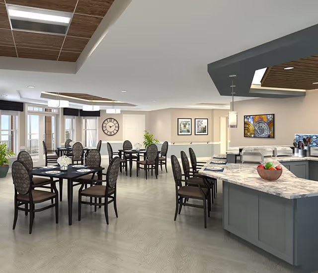 A spacious dining area in a senior living facility with multiple round tables and chairs arranged neatly. The room features large windows allowing natural light, a wall clock, framed artwork, and a kitchen counter with a bowl of fruit and place settings. The ceiling has recessed lighting and decorative wood panels.