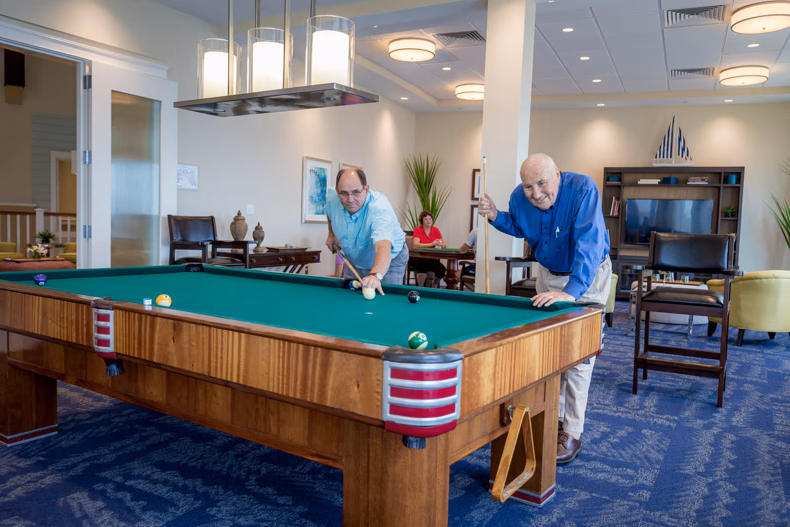 Two elderly men playing pool in a well-lit recreational room with blue carpet and wooden furniture. In the background, a woman is seated at a table, and there is a TV and decorative items on shelves.