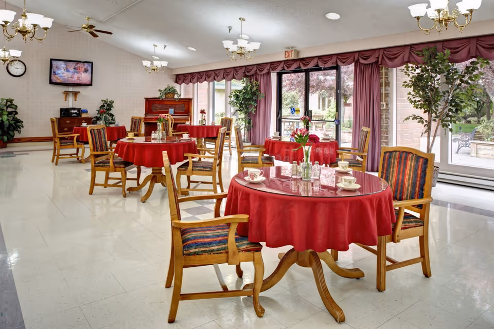 Bright communal dining room with round tables draped in red tablecloths, wooden chairs, chandeliers, and large windows overlooking a courtyard.