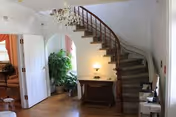 Interior view of a living area featuring a curved wooden staircase with carpeted steps, a small wooden table with a lamp underneath the stairs, a chandelier hanging from the ceiling, and an open doorway leading to another room with a large green plant visible.