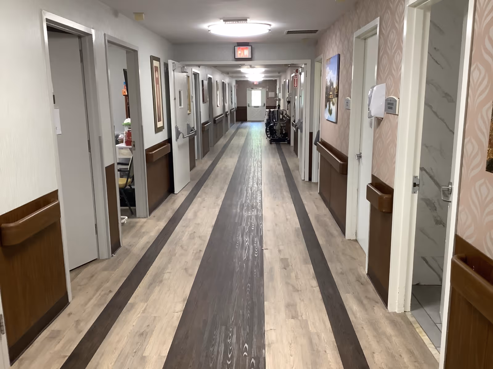 A long, well-lit hallway in a care facility with wood-patterned flooring and handrails on both sides. Several doors line the corridor, some open, revealing rooms and a bathroom with marble-patterned walls. Artwork and safety equipment are mounted on the walls, and an exit sign is visible at the far end.