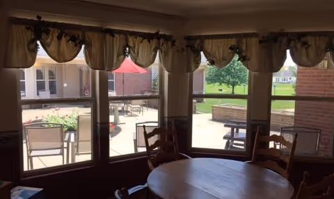 View from inside a room with a round wooden table and chairs, looking out through large windows with valance curtains onto an outdoor patio area with tables, chairs, a red umbrella, and greenery in the background.
