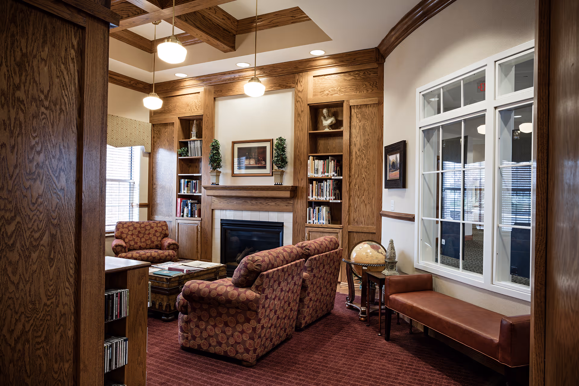 A cozy living room area with patterned armchairs and a loveseat arranged around a wooden coffee table. The room features a fireplace with built-in wooden bookshelves on either side, decorated with books, plants, and a bust sculpture. There is a large window with white framing on the right wall, a brown leather bench below it, and warm lighting from ceiling fixtures. The carpet is a deep red color, complementing the wood paneling and furniture.