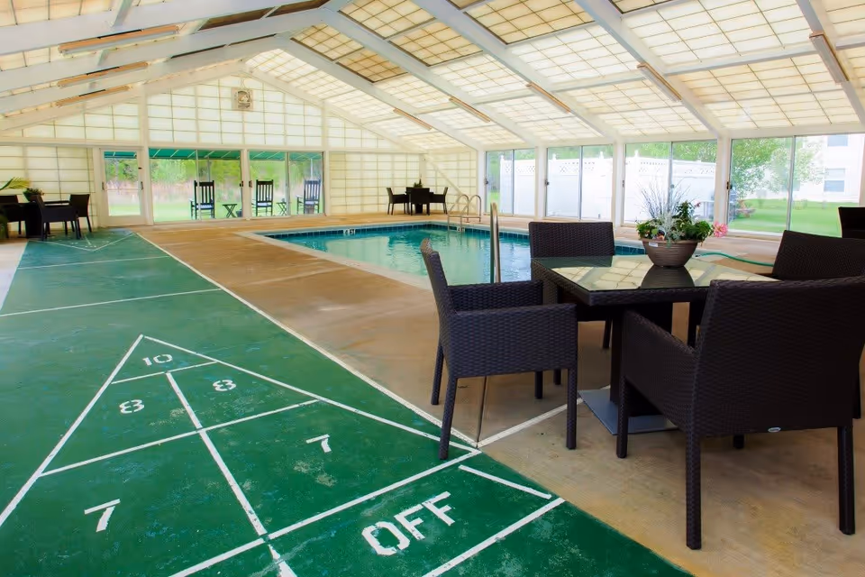 Indoor recreational area with a swimming pool, shuffleboard court, and several dark wicker chairs around glass tables. The space has a high, white paneled ceiling and large windows letting in natural light, with a view of greenery outside.