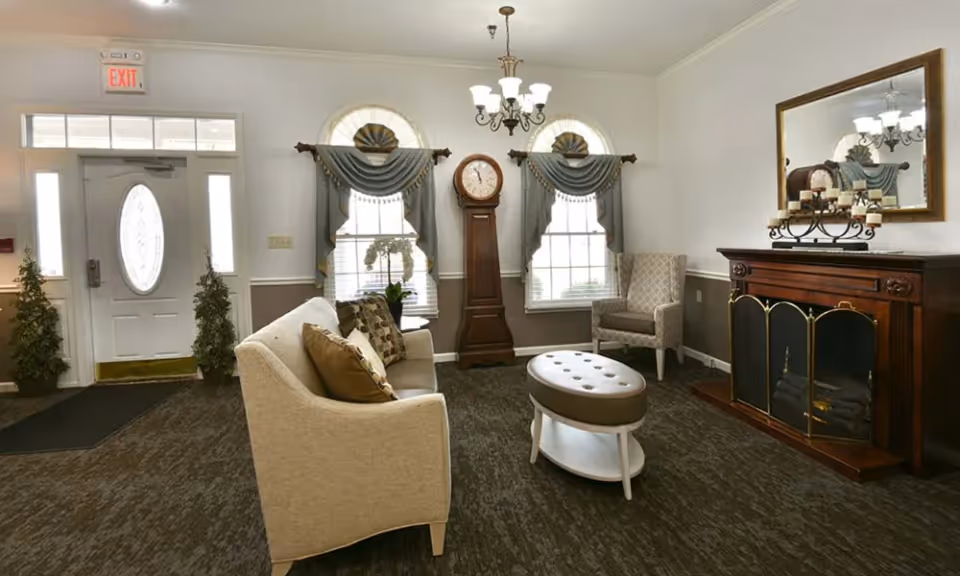 A cozy living room area with a beige sofa adorned with patterned pillows, a cushioned oval ottoman, and a single armchair. The room features two windows with decorative valances, a tall grandfather clock between the windows, a wooden fireplace with a mirror above it, and a chandelier hanging from the ceiling. The entrance door with an exit sign above is visible on the left side, along with two small potted plants.