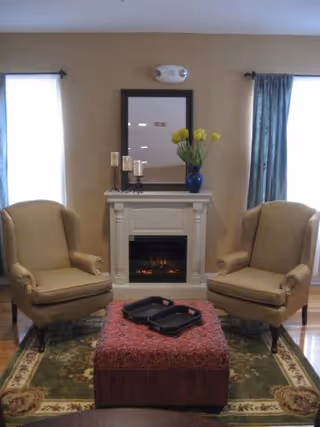 A cozy living room area with two beige upholstered armchairs facing each other, a red patterned ottoman in the center with two black trays on top, a green and beige floral area rug underneath, and a white fireplace with a mirror above it and a blue vase with yellow flowers on the mantel. There are two windows with curtains on either side of the fireplace.