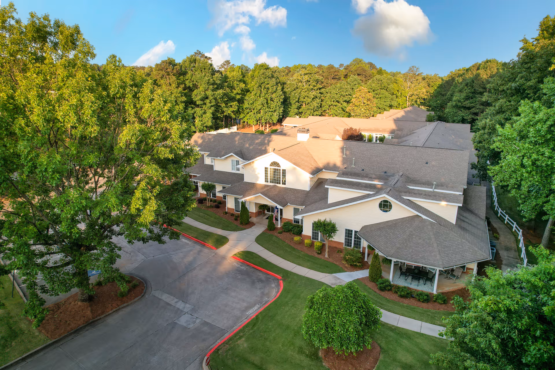 Aerial view of Legacy Ridge at Peechtree, showing a large senior living facility building surrounded by lush green trees and well-maintained lawns. The building has a beige exterior with multiple roof sections and a covered patio area with outdoor seating. The driveway and sidewalks are visible, along with a stop sign at the entrance.