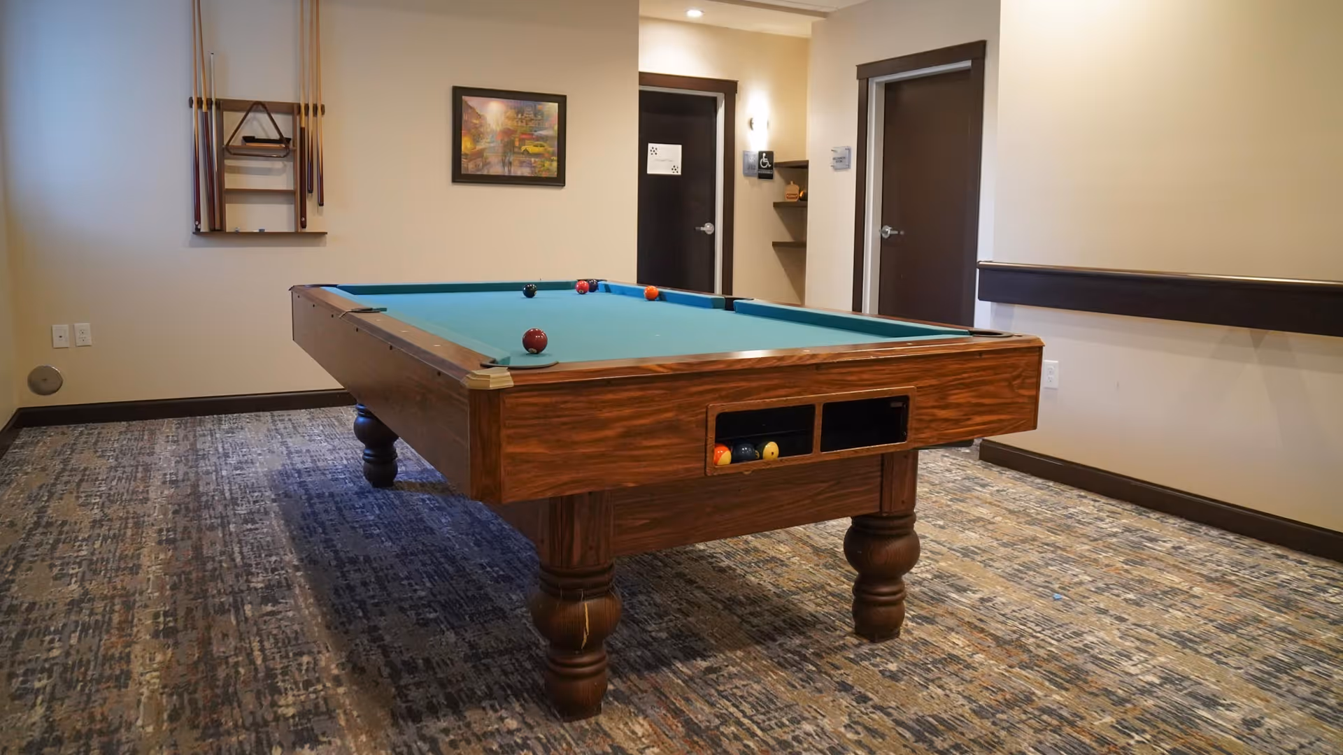 A room with a pool table in the center, featuring a blue felt surface and several pool balls on it. The room has patterned carpet flooring, beige walls, and two closed dark brown doors. A wall-mounted rack holds pool cues and a triangle rack. There is a framed picture on the wall and a small shelf near one of the doors.