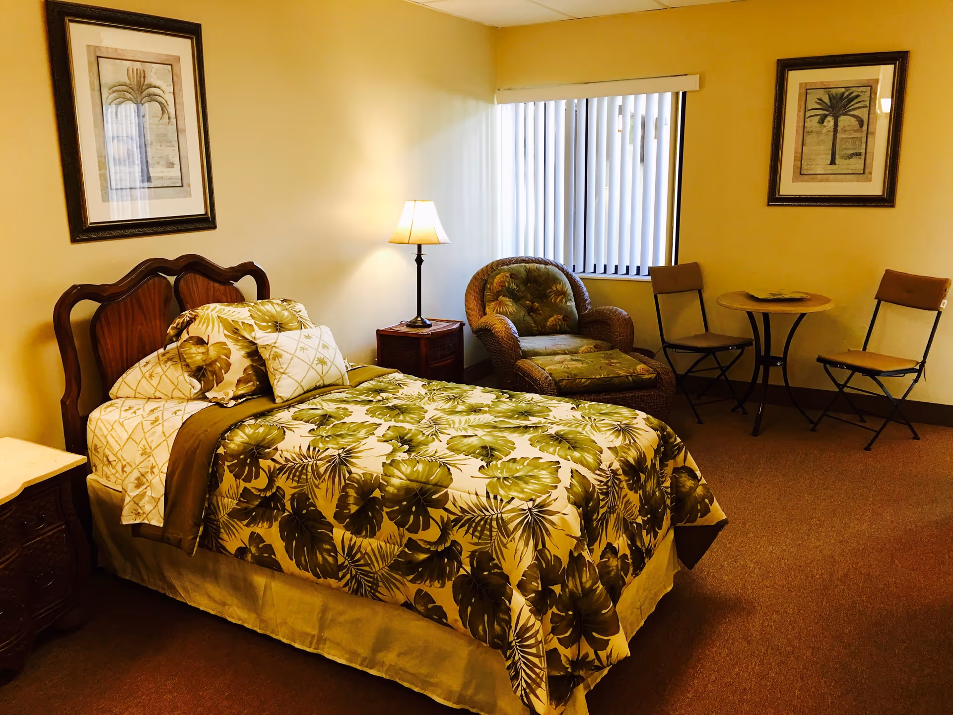 A cozy bedroom with a single bed featuring green and beige leaf-patterned bedding. There is a wooden headboard, a nightstand with a lamp, a cushioned wicker armchair with matching footrest, and a small round table with two chairs near a window with vertical blinds. Two framed palm tree artworks hang on the walls.