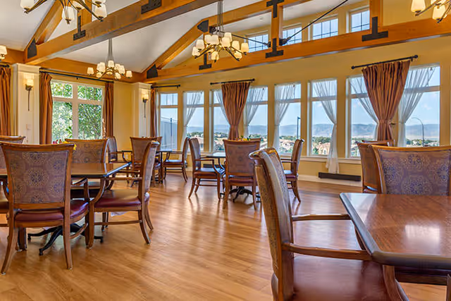 A spacious dining room with wooden floors, multiple tables and chairs, large windows with sheer curtains, and a vaulted ceiling with exposed wooden beams and chandeliers. The windows offer a scenic view of mountains and greenery outside.