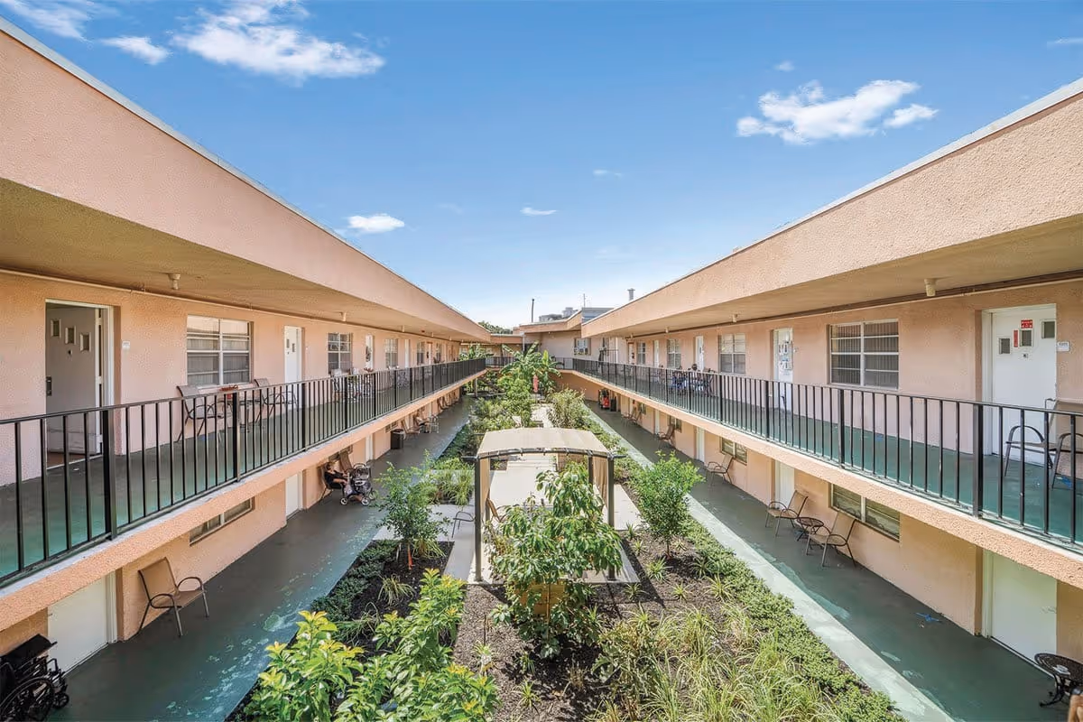 View of an outdoor courtyard area in a senior living community with two levels of walkways lined with railings and doors to individual units. The courtyard features a garden with various plants and a shaded seating area in the center. The building exterior is light pink with green flooring on the walkways, under a clear blue sky.
