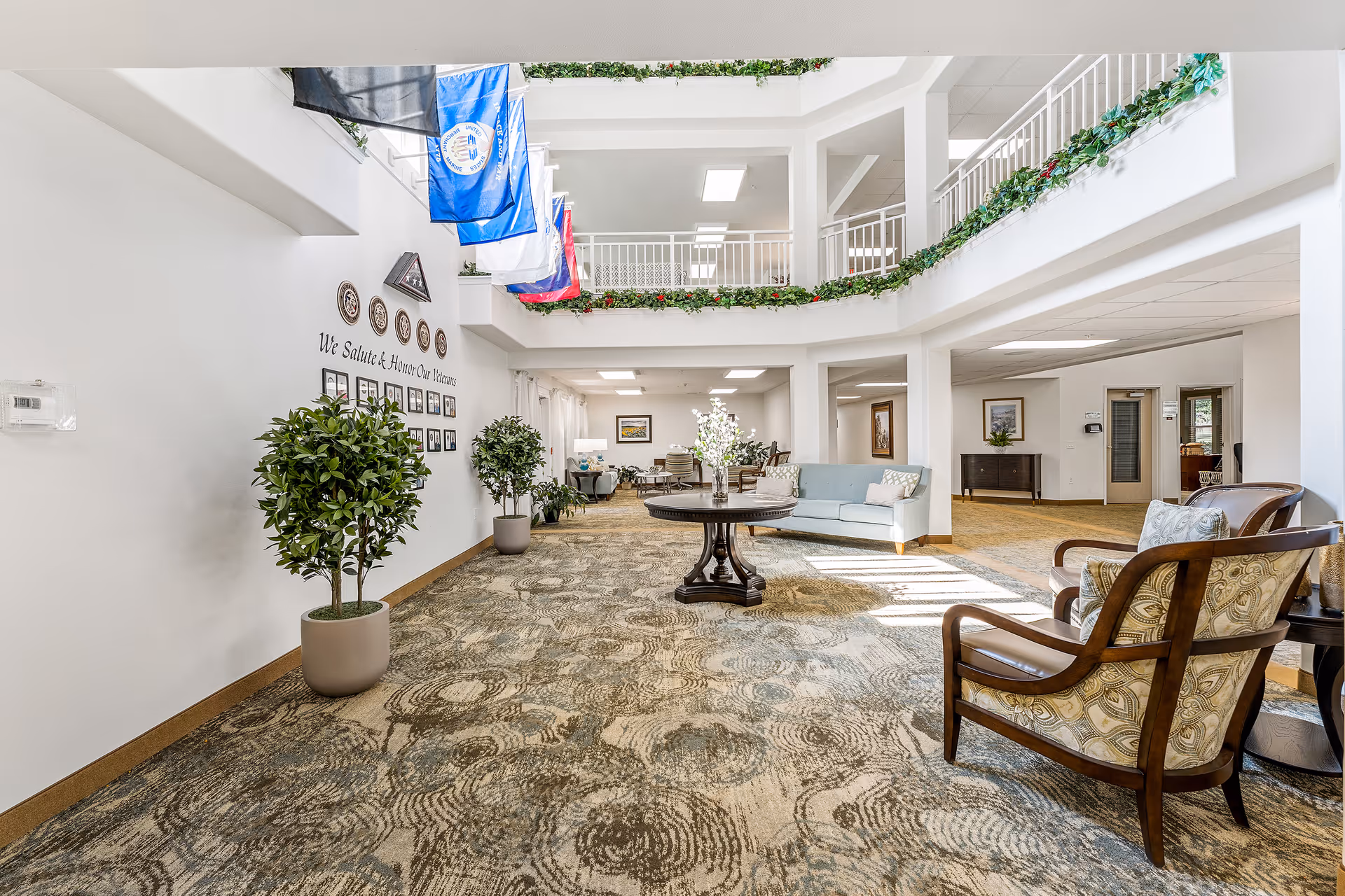 Bright and spacious interior common area with patterned carpet, potted plants, a round wooden table with a floral arrangement, upholstered chairs, and a light blue sofa. The area features a high ceiling with a second-floor balcony decorated with green garlands and several hanging flags. Wall plaques and framed photos are displayed on one wall.