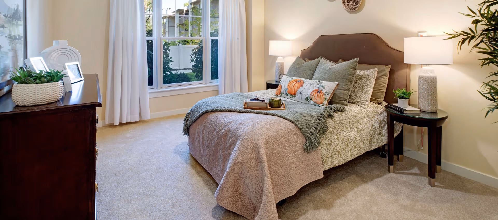 A cozy bedroom with a neatly made bed featuring a beige quilt, green throw blanket, and decorative pillows including one with a pumpkin design. There are two bedside tables with lamps and small plants, a dark wooden dresser with framed photos and a plant, beige carpet, and a large window with white curtains letting in natural light.