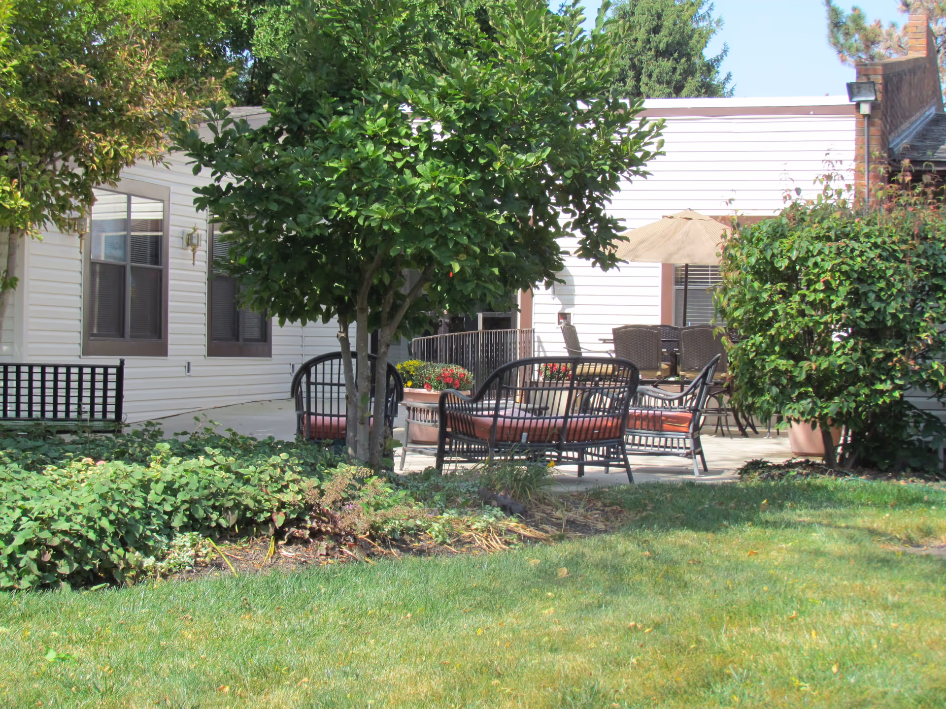 Outdoor patio area at Springfield Assisted Living with metal chairs and benches with cushions, a table with an umbrella, surrounded by green bushes, trees, and grass.