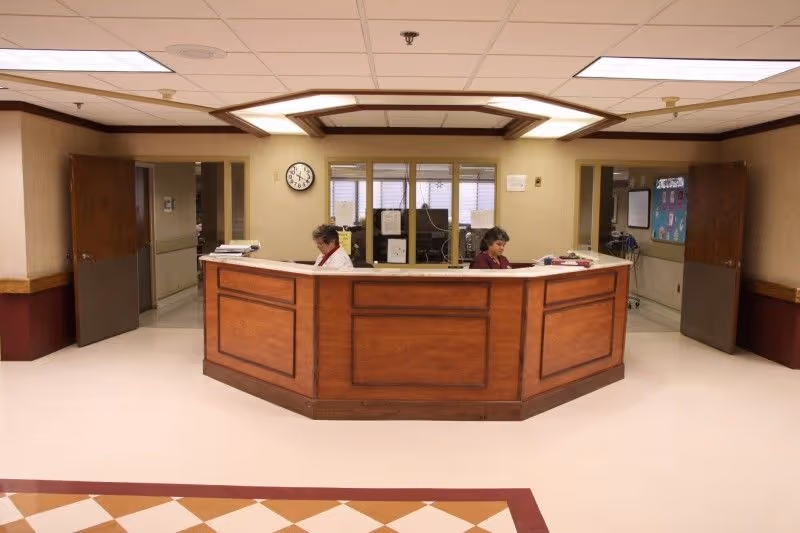 Reception area in a senior living facility with a wooden front desk where two staff members are seated working. The area has beige walls, a clock on the wall, open doors on both sides, and a tiled floor with a patterned border.