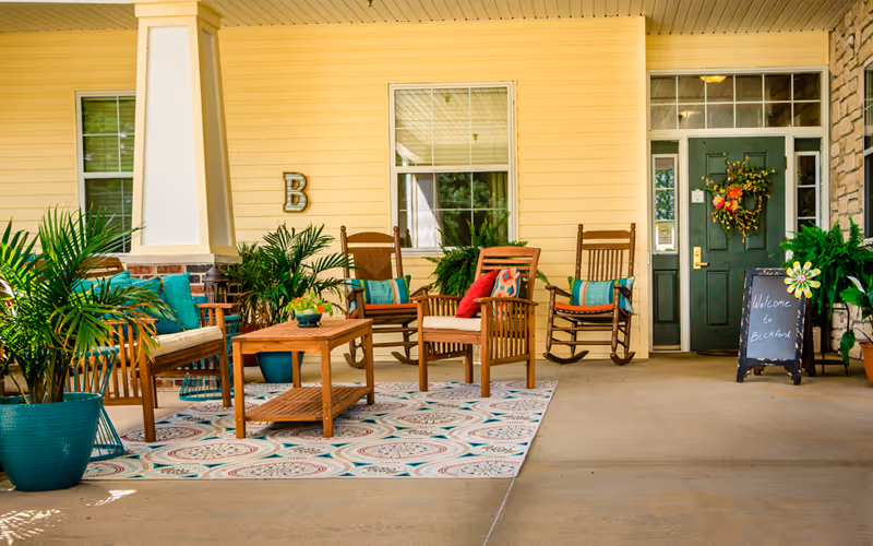 Covered outdoor seating area with wooden chairs and a table on a patterned rug, surrounded by potted plants. A green door with a wreath and a chalkboard sign that says 'Welcome to Bickford' are visible on the right side.