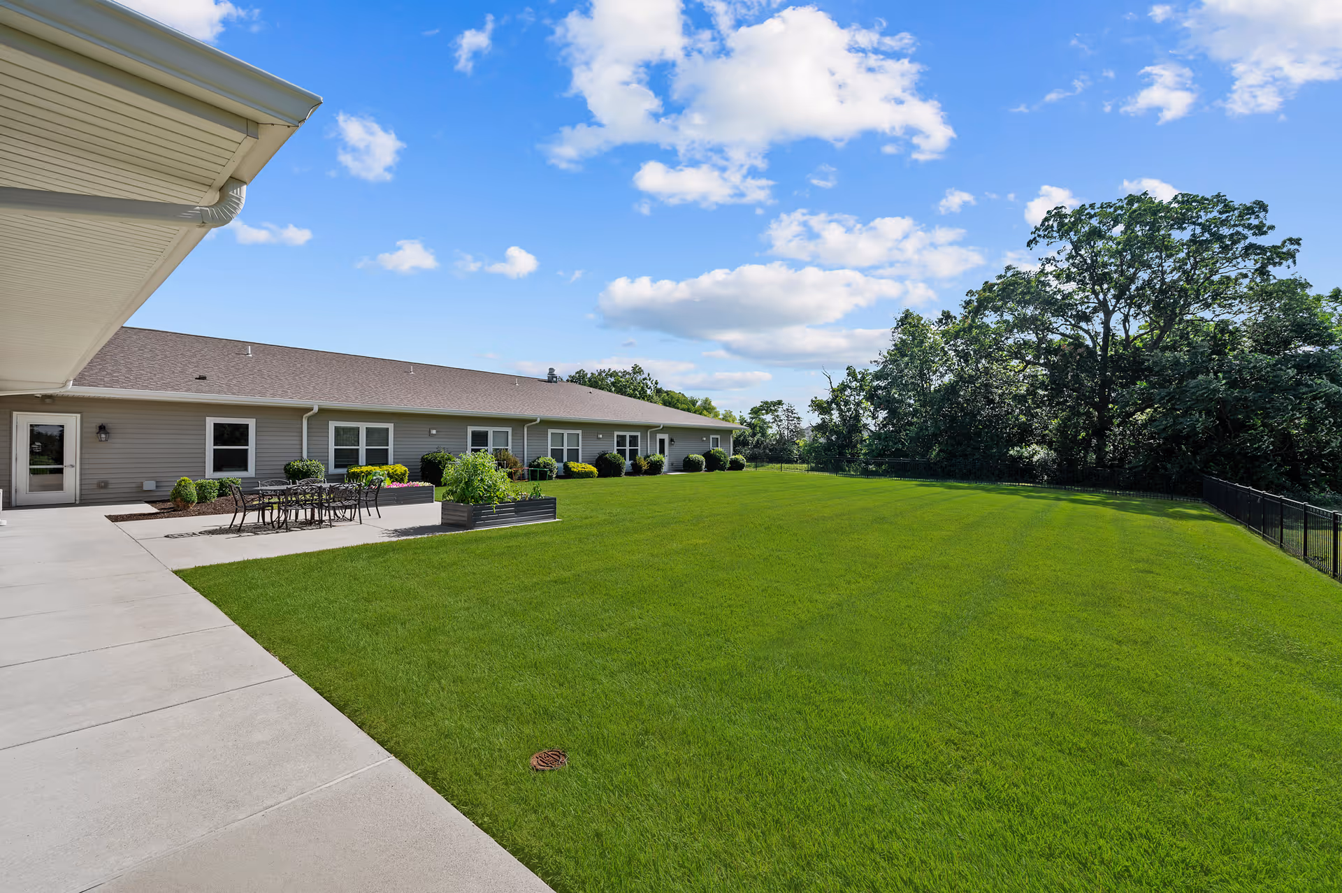 Outdoor view of a senior living facility showing a large green lawn bordered by a concrete walkway and a single-story building with multiple windows and a patio area with outdoor furniture. Trees and a blue sky with clouds are visible in the background.