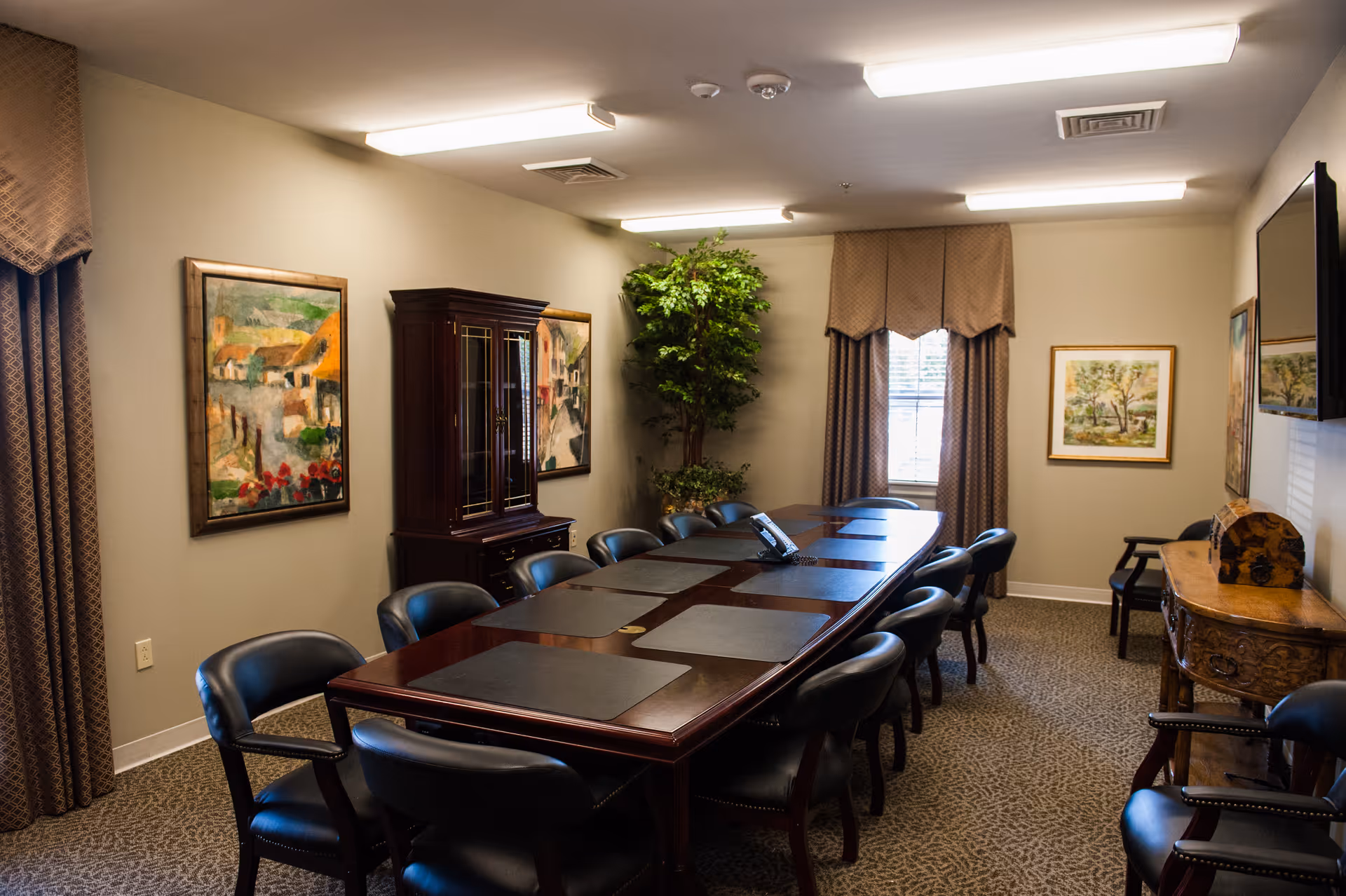 A conference room with a long wooden table surrounded by black leather chairs. The room has beige walls adorned with framed paintings and a large window with curtains. There is a wooden cabinet and a decorative plant in the corner, and a telephone is placed on the table.