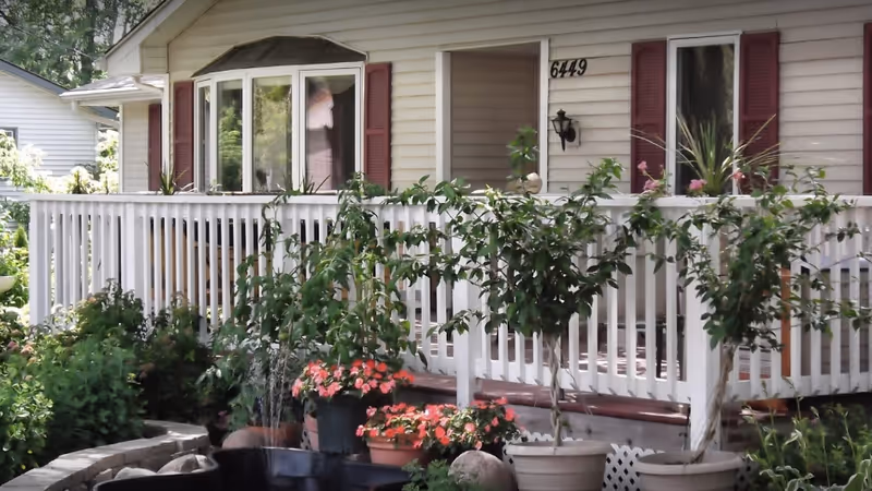 Front porch of a light-colored house with white railing, potted plants, and greenery in the garden. The house has maroon shutters and the number 6449 is visible near the entrance.