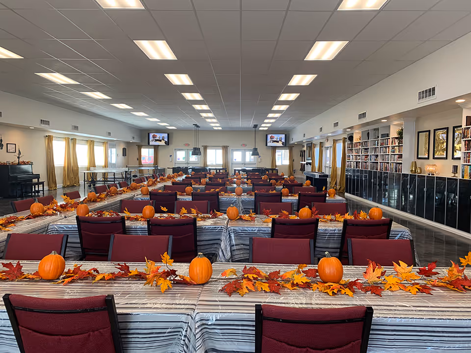 A large dining room decorated for autumn with long tables covered in striped tablecloths, each adorned with small pumpkins and colorful fall leaves. The room has maroon chairs, large windows with yellow curtains, bookshelves along one wall, and multiple ceiling lights. Two televisions are mounted on the far wall.