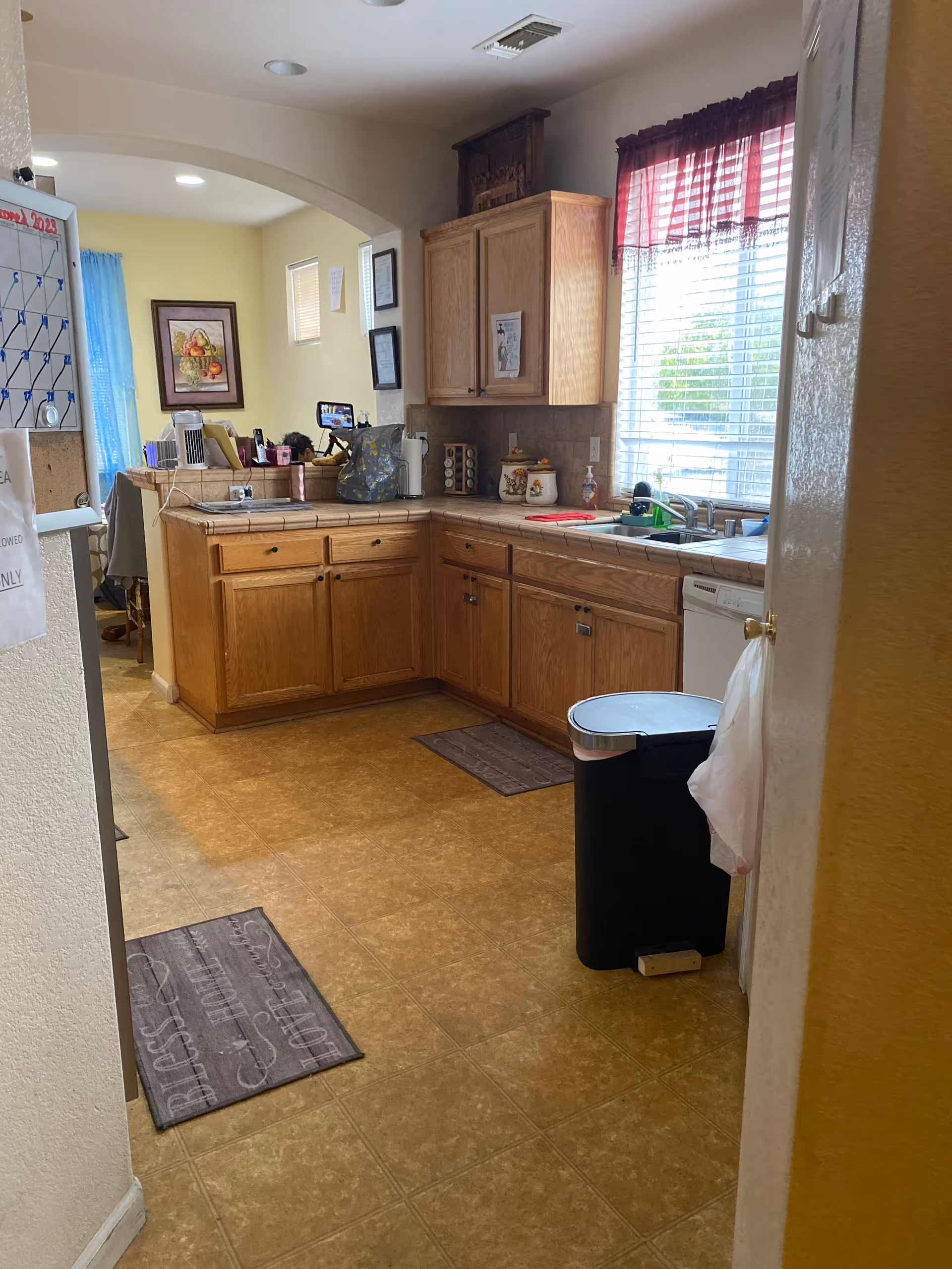 Interior view of a kitchen with wooden cabinets, a countertop with various items including a bag and containers, a window with blinds and a red valance, a trash can with a plastic bag hanging on the side, and a tiled floor with two mats. The kitchen opens into another room with a desk and chair visible.