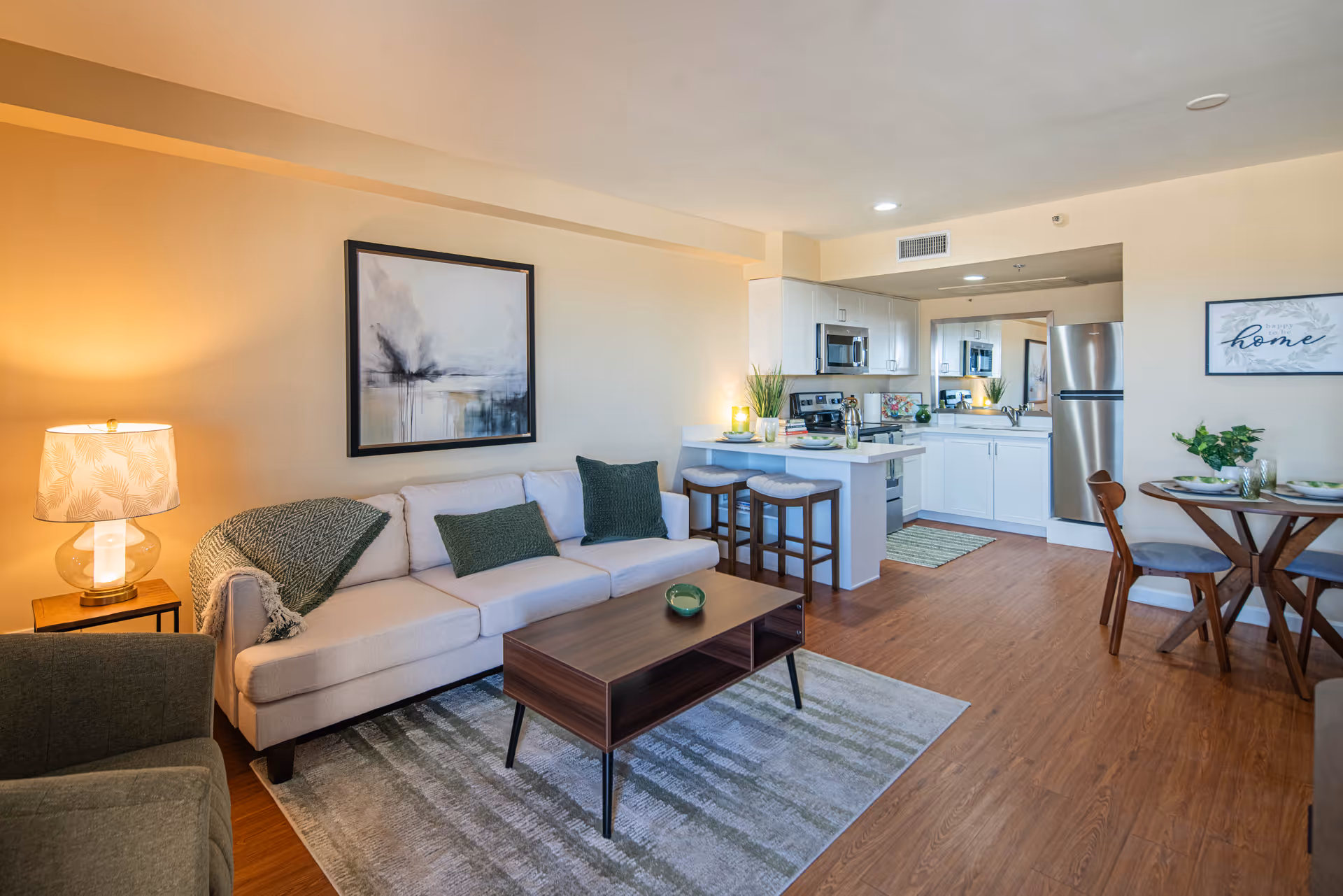 A bright and modern open-concept living area featuring a beige sofa with green cushions, a wooden coffee table on a patterned rug, a side table with a lamp, and a kitchen area with white cabinets, stainless steel appliances, and a breakfast bar with stools. A small dining table with chairs is visible on the right, along with wall art and decorative plants.