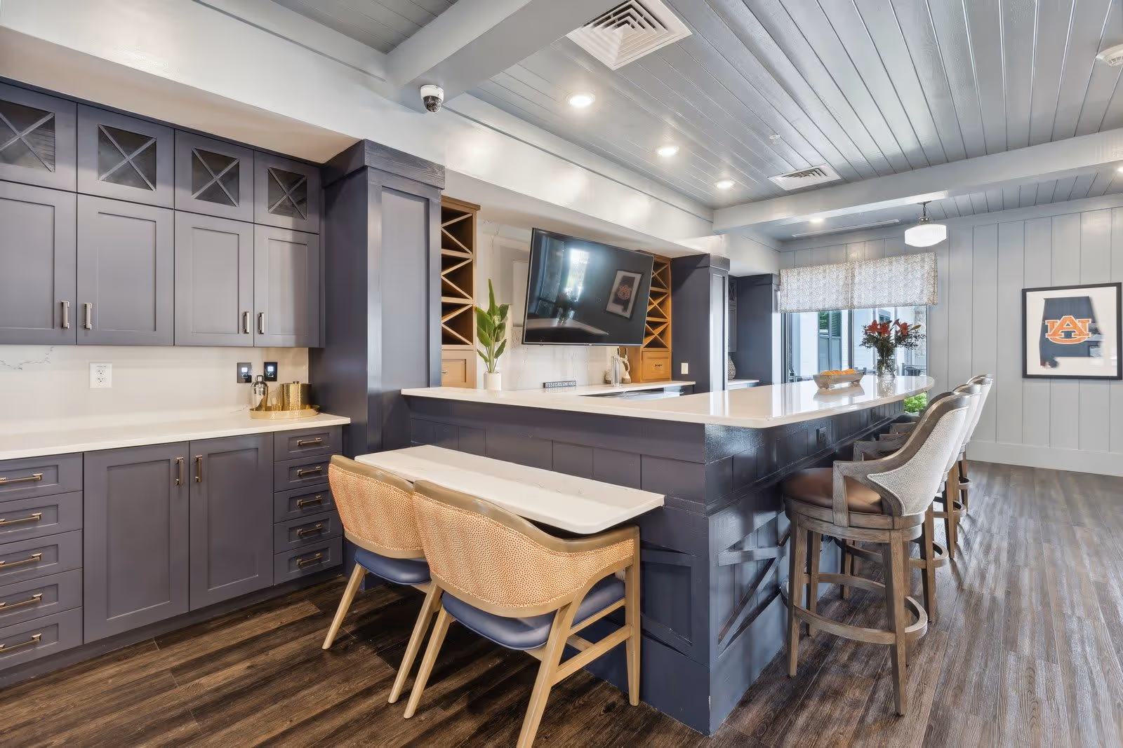 Modern kitchen area with dark gray cabinetry, a large white countertop island with bar stools, a mounted flat-screen TV, and a small table with two chairs. The room has wood flooring, recessed lighting, and a window with a patterned shade. A framed Auburn University logo is visible on the wall.