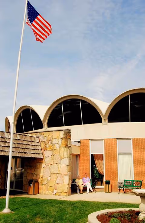 Exterior view of Envive Healthcare of Indianapolis with flag, arched windows, and two residents seated.