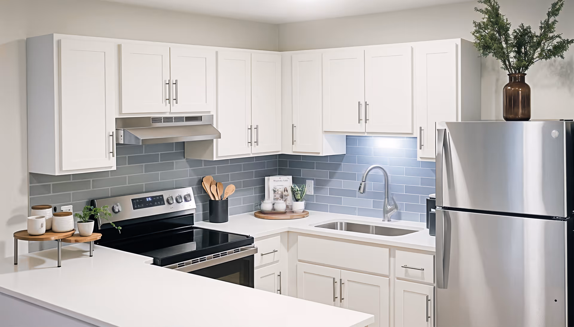 Modern kitchen with white cabinets, stainless steel refrigerator and oven, gray subway tile backsplash, a sink with a curved faucet, and decorative items including plants and wooden utensils.