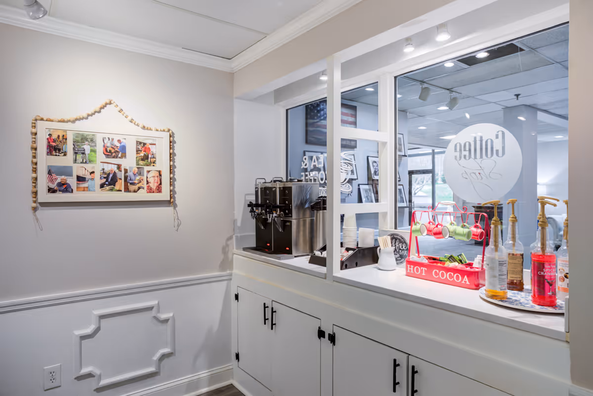 A coffee and hot cocoa station in a senior living facility with a stainless steel coffee machine, cups, stirrers, and various syrup bottles. There is a window with the words 'Coffee Stop' visible, and a framed collage of photos on the adjacent wall. The area is clean and well-lit with white cabinetry below the counter.
