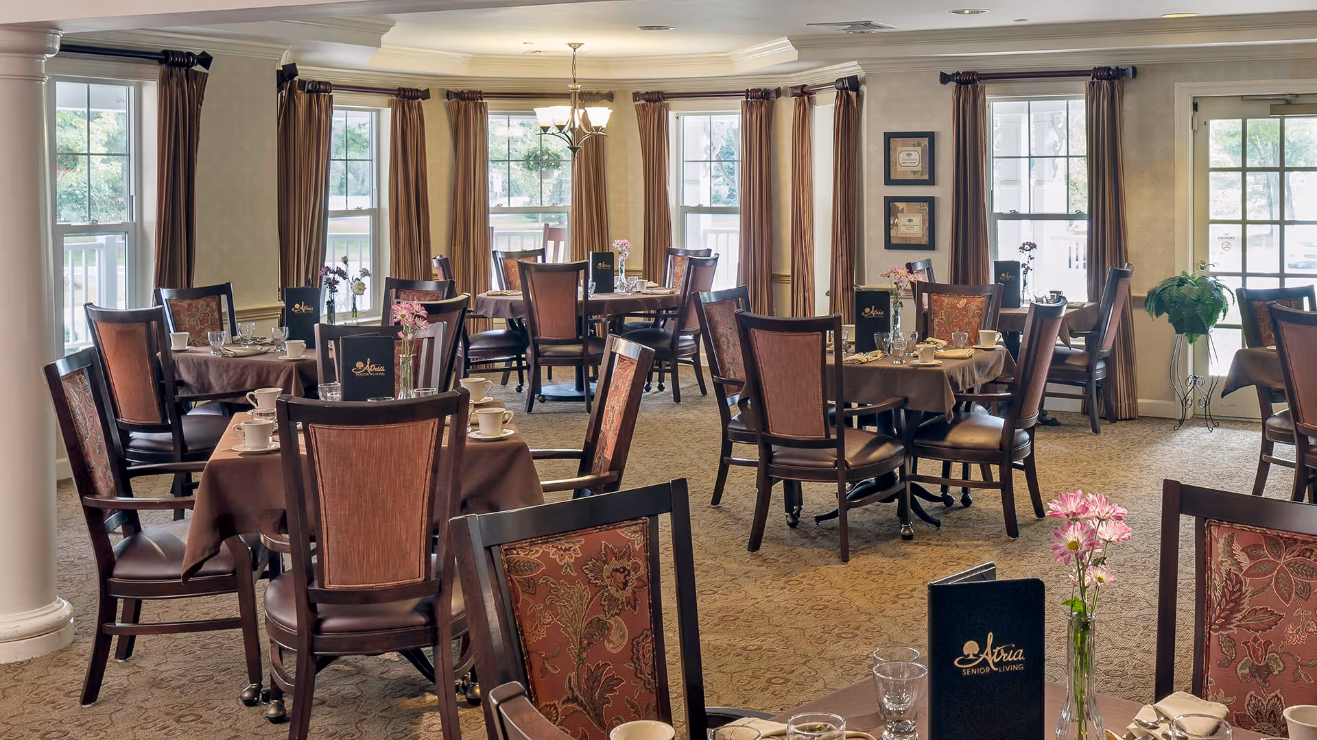 A well-lit dining room with multiple round tables covered in brown tablecloths, each set with cups, glasses, and menus labeled 'Atria Senior Living'. The room features large windows with beige curtains, decorative plants, and framed artwork on the walls.