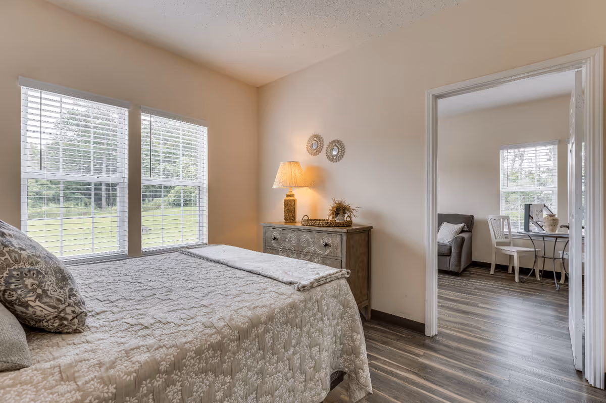 A cozy bedroom with a bed covered in a patterned quilt and pillows, two large windows with white blinds letting in natural light, a wooden dresser with a lit table lamp and decorative items, and an open doorway leading to a room with a chair, table, and windows.