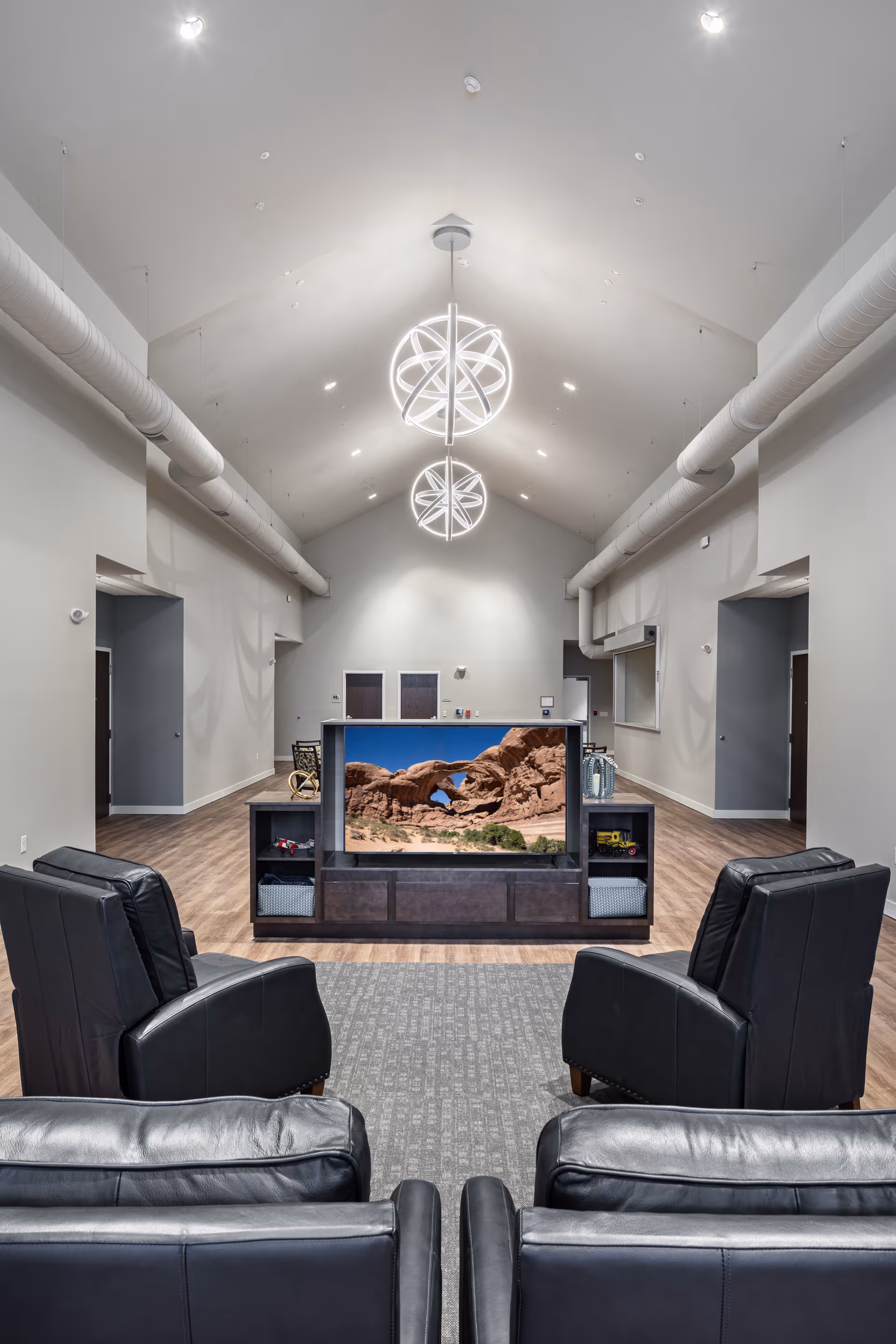 Spacious common living area with a vaulted ceiling and modern pendant lights, four black leather recliners facing a TV console showing a desert landscape.