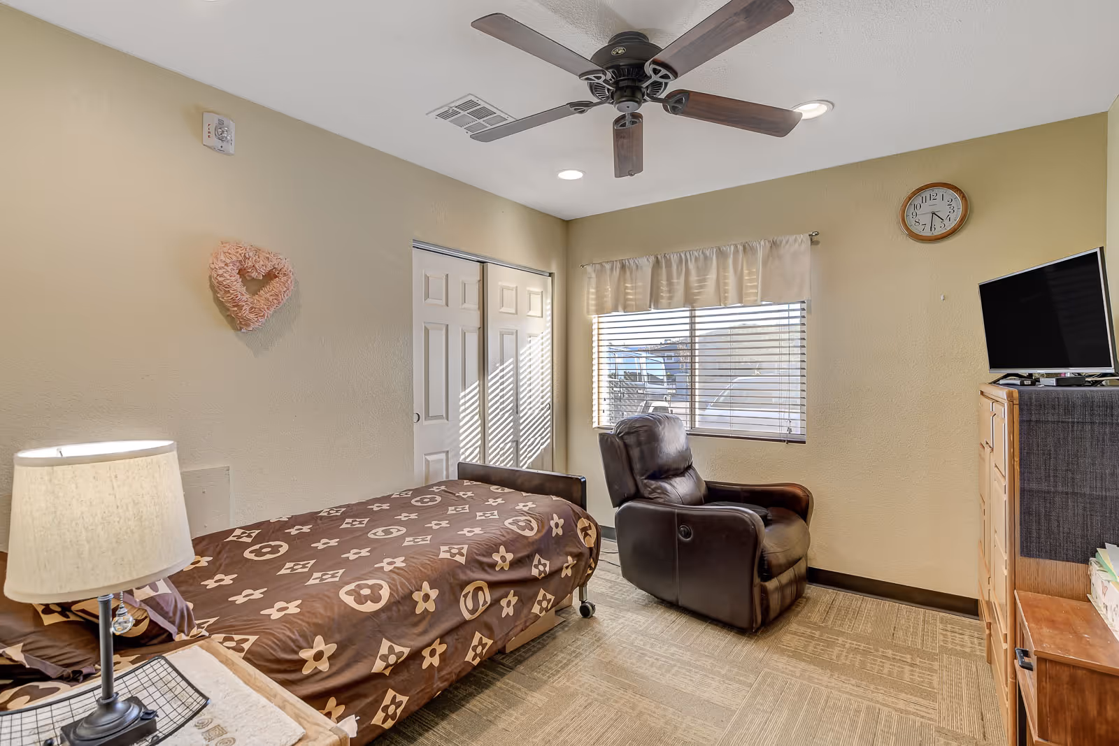 A cozy bedroom with a single bed covered in a brown patterned blanket, a bedside table with a lamp, a brown leather recliner chair near a window with blinds and a valance, a wooden dresser with a TV on top, a wall clock, and a ceiling fan with wooden blades.