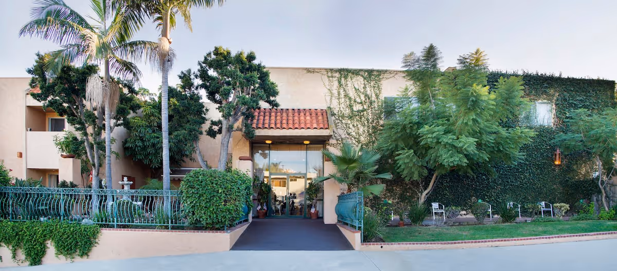 Front exterior view of Palos Verdes Villa facility showing an entrance with glass doors, surrounded by lush greenery including palm trees, bushes, and ivy-covered walls under a clear sky.