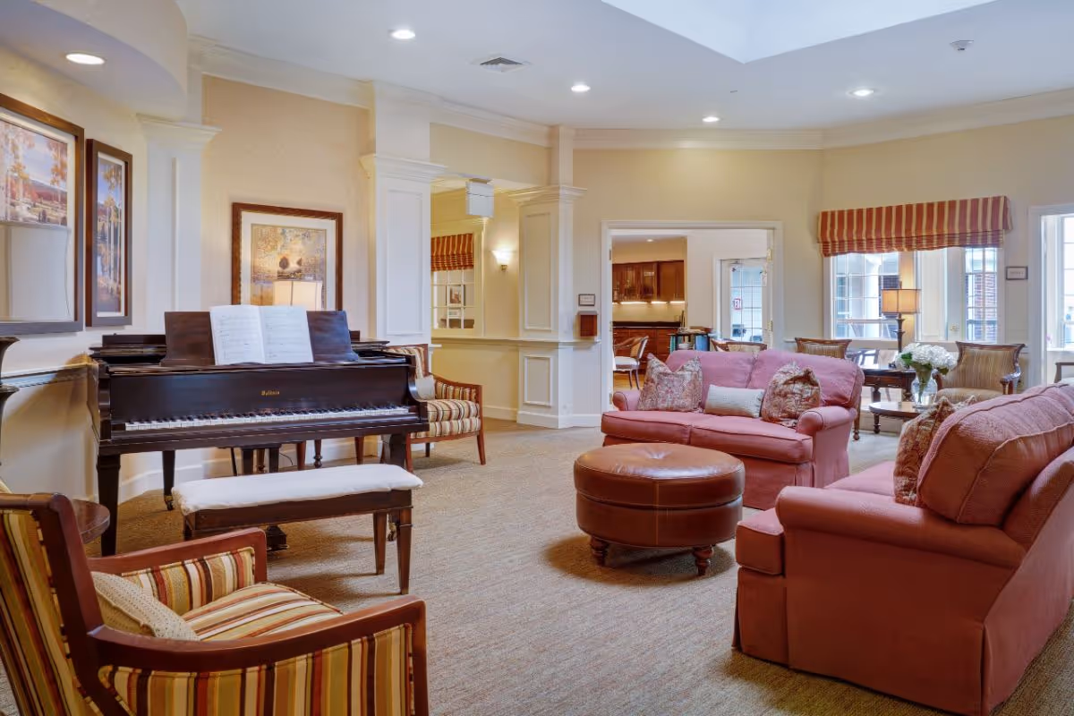 A bright communal lounge featuring a grand piano, upholstered sofas and chairs arranged around a round leather ottoman.