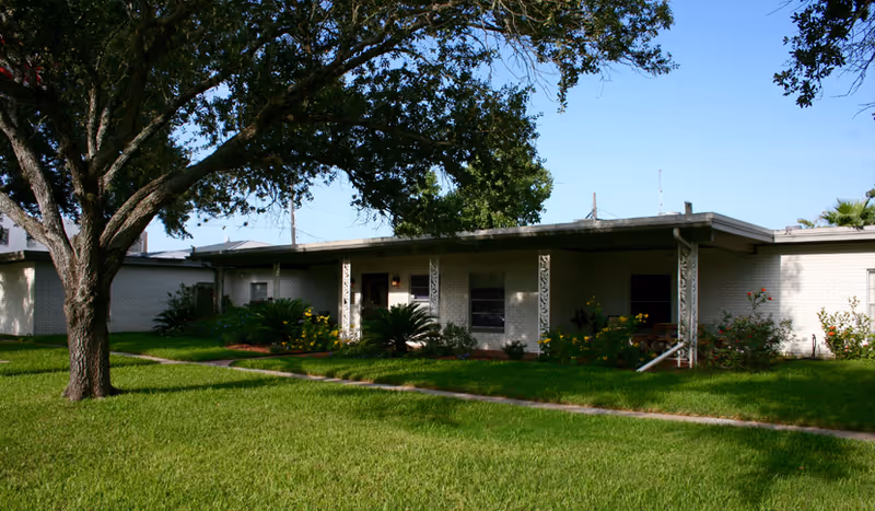 Single-story nursing and rehab center building with a flat roof, white brick exterior, and decorative metal columns on the porch. The building is surrounded by a well-maintained lawn, shrubs, flowers, and a large tree providing shade in the foreground under a clear blue sky.