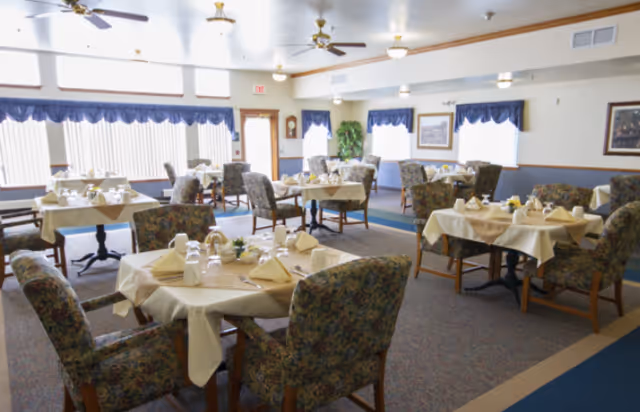 Bright dining room with multiple round tables set with napkins, glassware, and upholstered chairs.