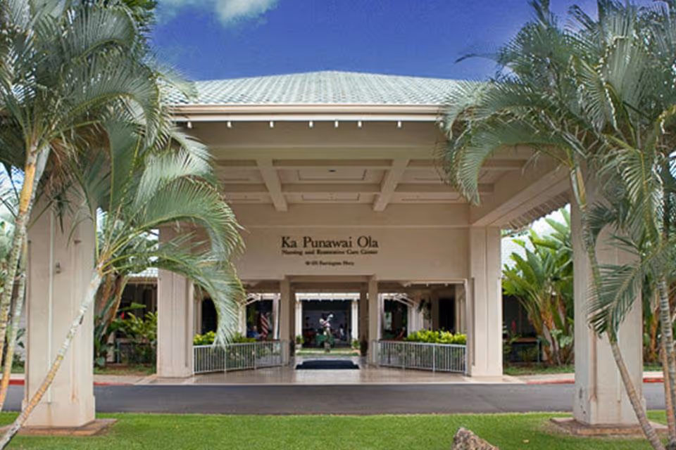Entrance of Ka Punawai Ola Nursing and Restorative Care Center with a covered driveway, surrounded by palm trees and greenery under a blue sky with some clouds.