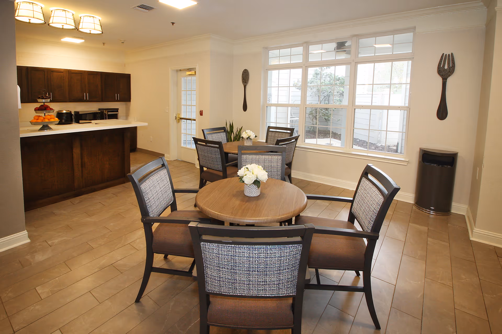 A dining area in a senior living facility featuring two round wooden tables each surrounded by four cushioned chairs with patterned backs. The room has large windows letting in natural light, a kitchen counter with dark wood cabinets, and decorative oversized spoon and fork wall hangings. A trash can is placed in the corner near the window.