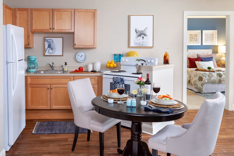Small open-plan kitchen with wooden cabinets, a white stove and fridge, and a round dining table set for two with a bedroom visible through a doorway.
