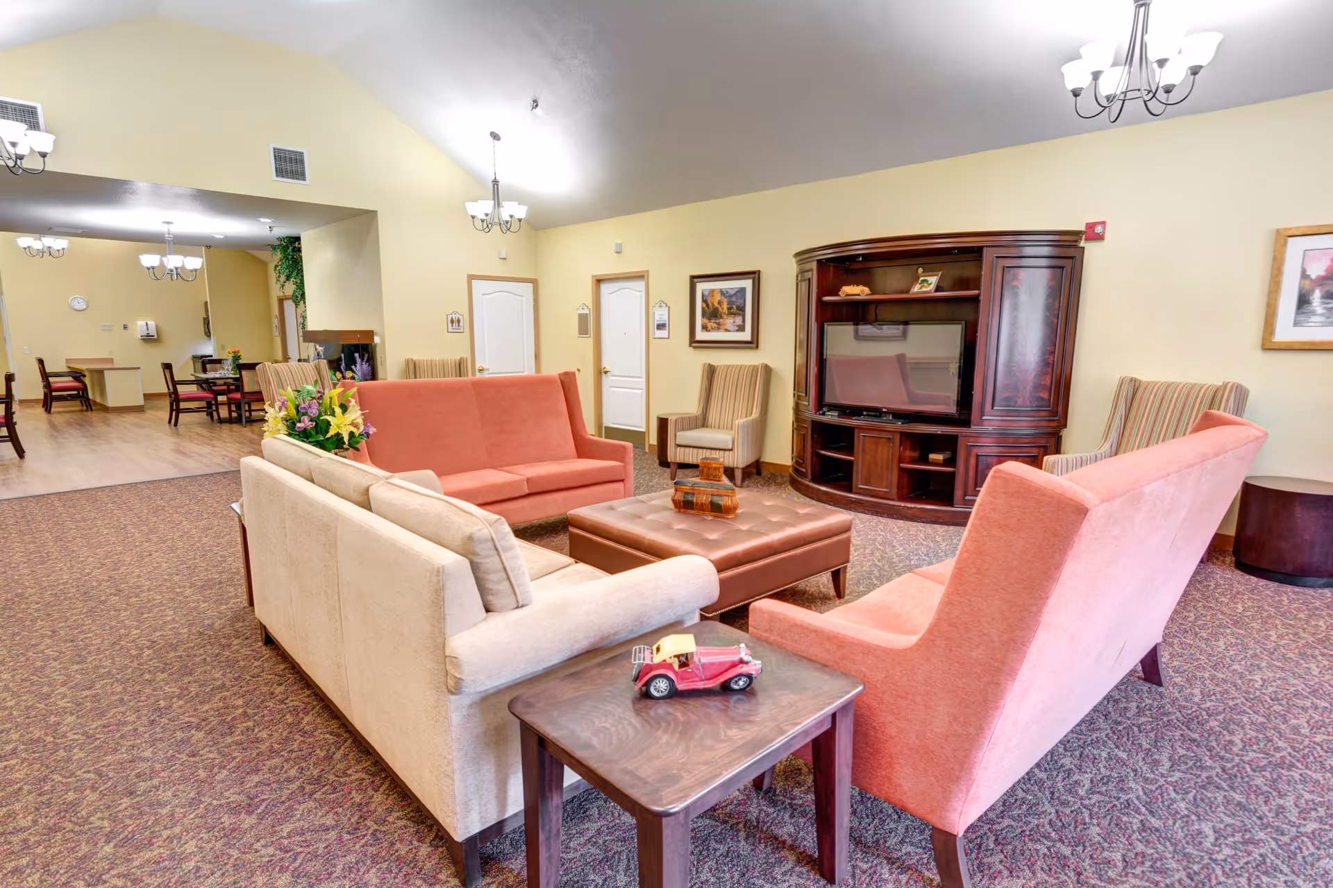 A cozy living room area in a senior living facility with beige and coral sofas arranged around a large brown ottoman. There is a wooden entertainment center with a TV, two armchairs, framed artwork on the walls, and a small side table with a decorative toy car. The room has carpeted flooring and soft yellow walls, with a dining area visible in the background.