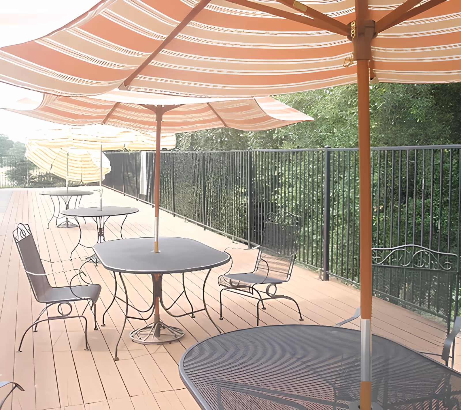 Outdoor patio area with multiple metal tables and chairs, each table shaded by large striped umbrellas. The patio is enclosed by a black metal fence and surrounded by greenery.