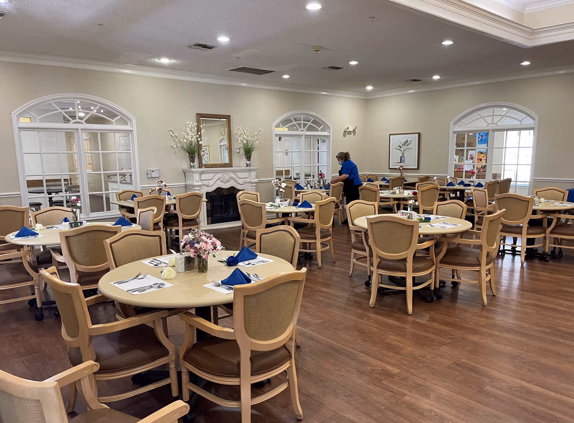 A spacious dining room with multiple round tables set with blue napkins, silverware, and small flower arrangements. The room has wooden floors, beige walls, and large windows with decorative white frames. A person wearing a blue shirt and a face mask is arranging items on one of the tables. There is a white decorative fireplace with a mirror and flower vases above it on one wall.