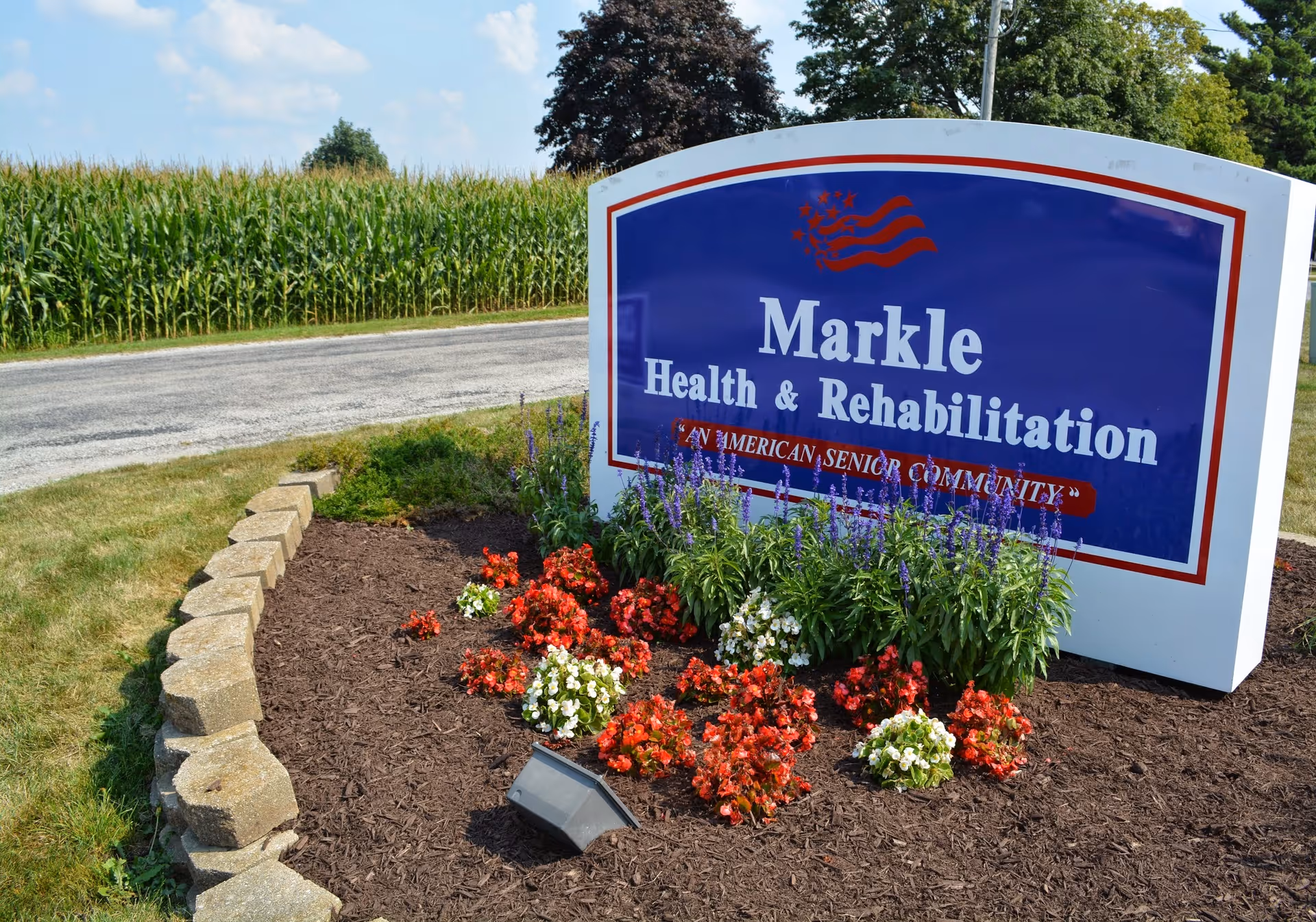 A large blue entrance sign reading "Markle Health & Rehabilitation" sits in a landscaped flowerbed beside a road and cornfield.