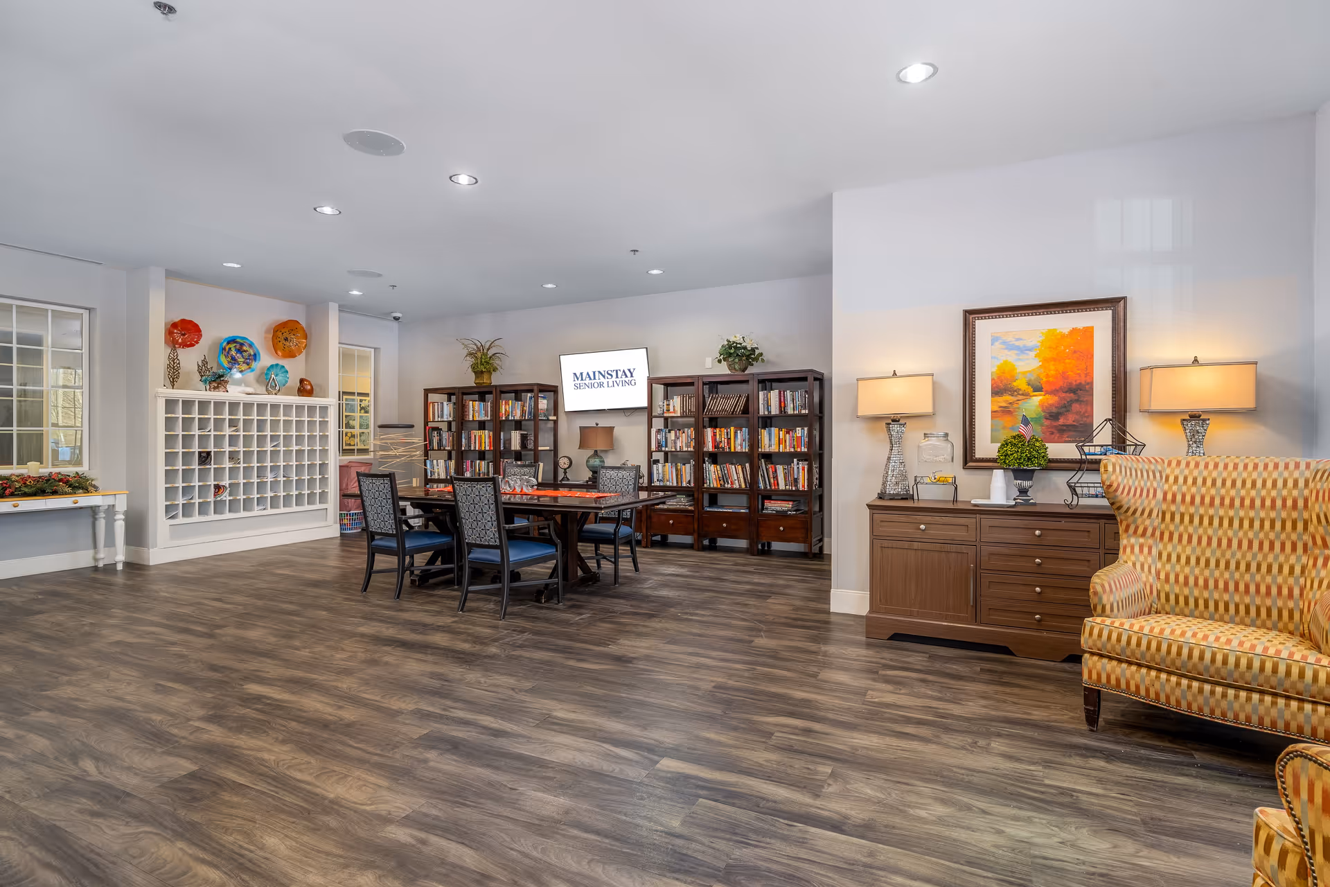 A spacious senior living common area with wooden flooring, a table with six chairs in the center, bookshelves filled with books against the back wall, and a TV screen displaying 'MAINSTAY SENIOR LIVING'. To the right, there is a wooden sideboard with two lamps, a decorative plant, and a framed painting of an autumn landscape above it. A patterned armchair is partially visible on the right side. The left side features a white cubby shelf with decorative plates and other items on top.