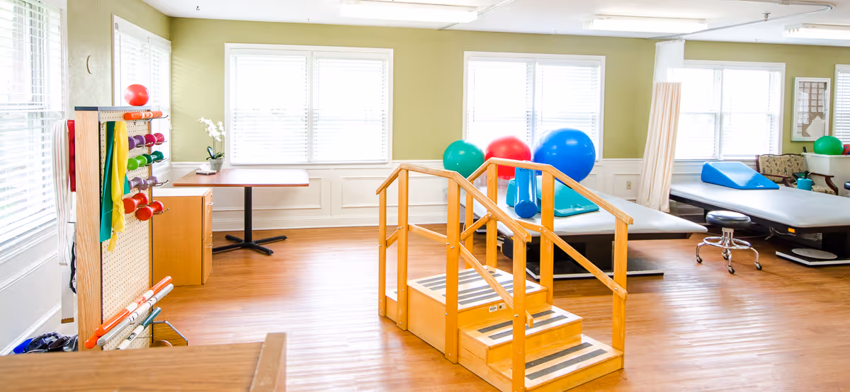 A bright physical therapy room with wooden flooring and light green walls. The room contains exercise equipment including colorful dumbbells on a rack, resistance bands, large exercise balls, therapy tables, and a wooden set of stairs with handrails for rehabilitation exercises. Large windows with white blinds allow natural light to fill the space.