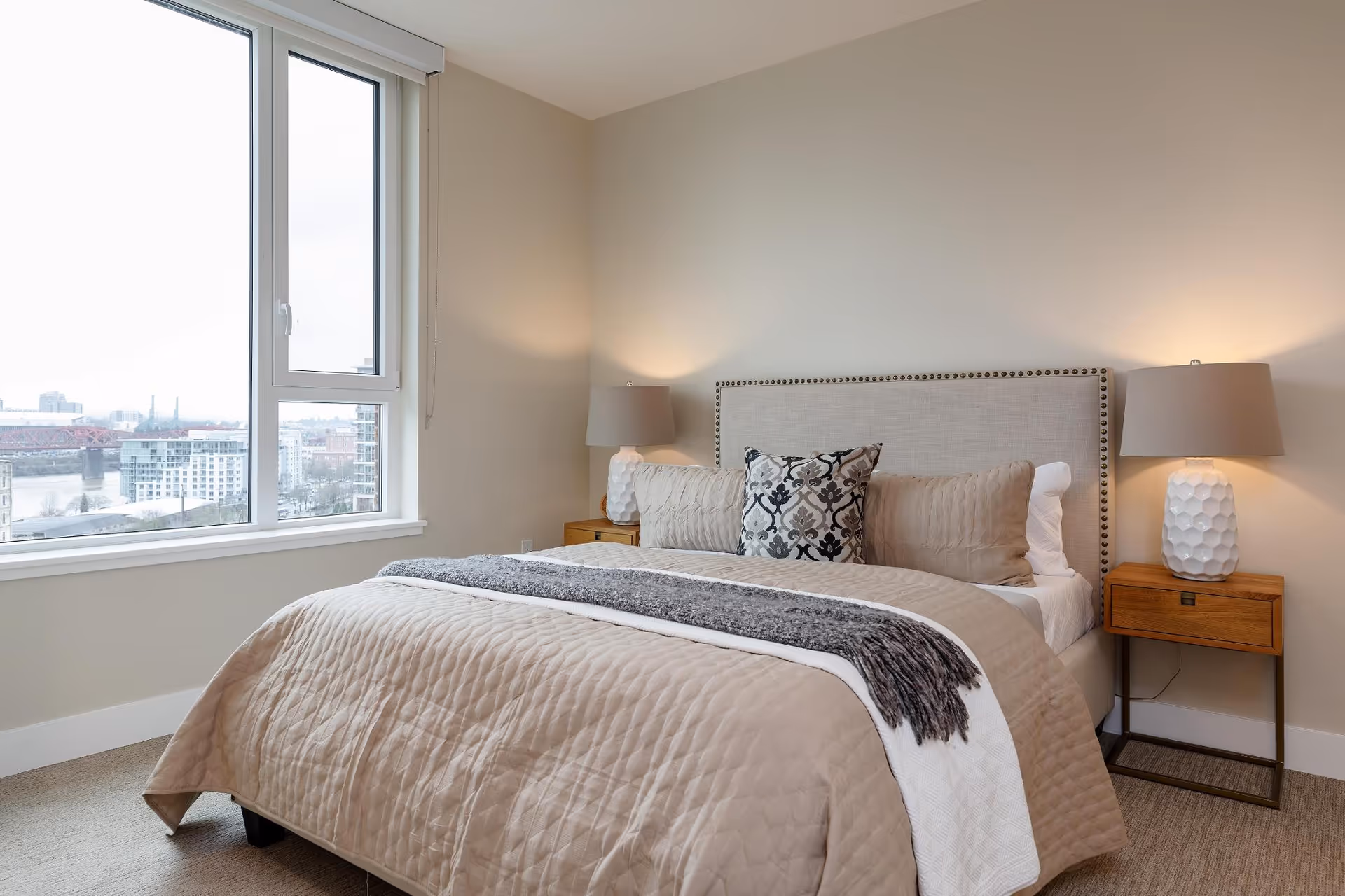 Well-lit bedroom with a bed dressed in neutral bedding, two bedside tables with lamps, and a large window overlooking the city.
