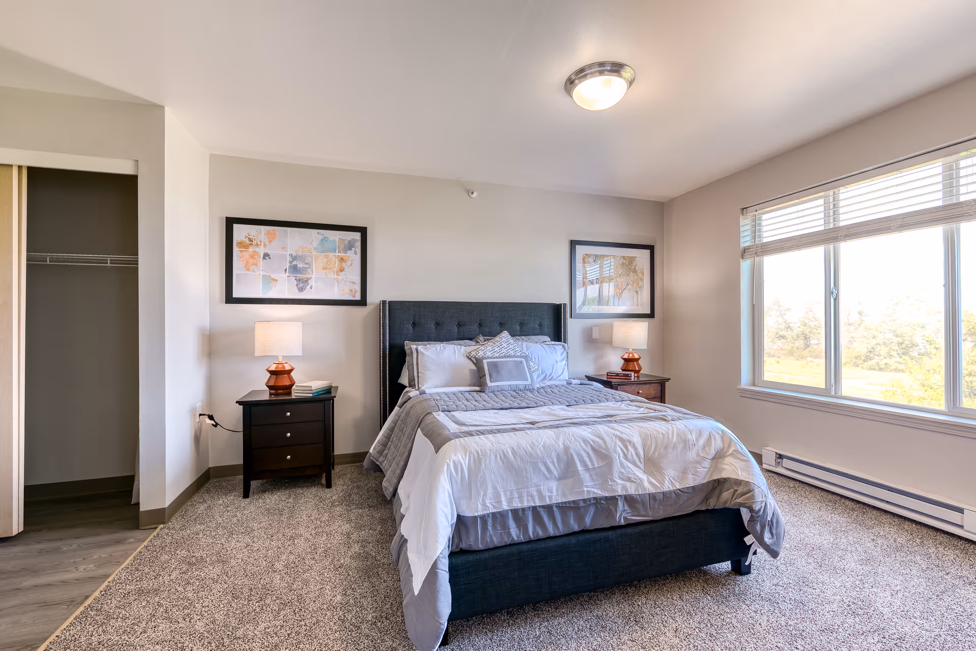 A bright and clean bedroom with a large window letting in natural light. The room features a neatly made bed with gray and white bedding, two bedside tables each with a lamp, framed artwork on the walls, and an open closet with sliding doors. The floor is carpeted near the bed and transitions to wood flooring near the closet.