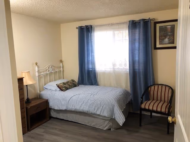 A small bedroom with a single bed dressed in blue check bedding, a nightstand and lamp, a striped chair, and blue curtains over a window.